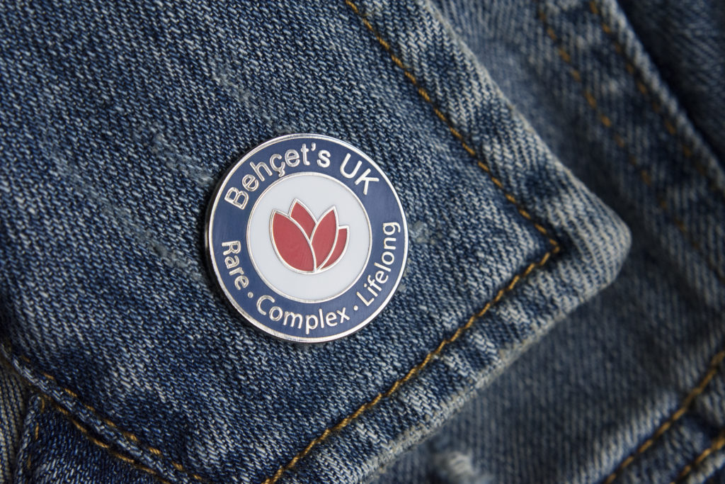 Close-up photo of a Behçet's UK round pin badge on a denim jacket. 