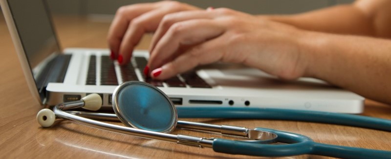 Photo shows hands typing on a laptop. There is a stethoscope next to the laptop.