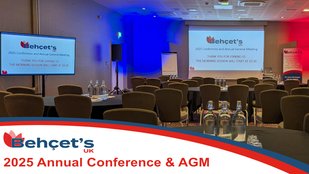 Conference room with chairs around tables for attendees and 2 large screens showing the Behçet’s UK logo. With the text which reads "Behçet's UK 2023 Annual Conference & AGM"