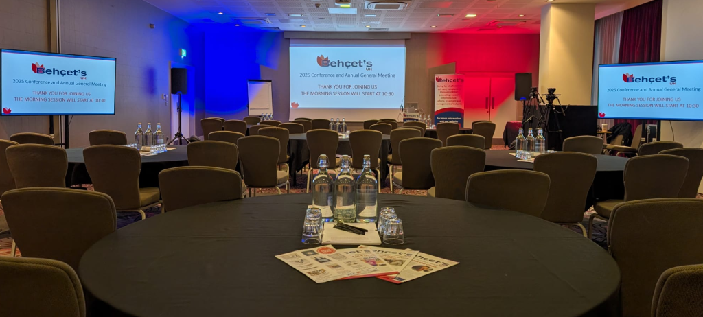 Conference room with chairs around tables for attendees and 3 large screens showing the Behçet’s UK logo, set up for the 2025 Conference and AGM