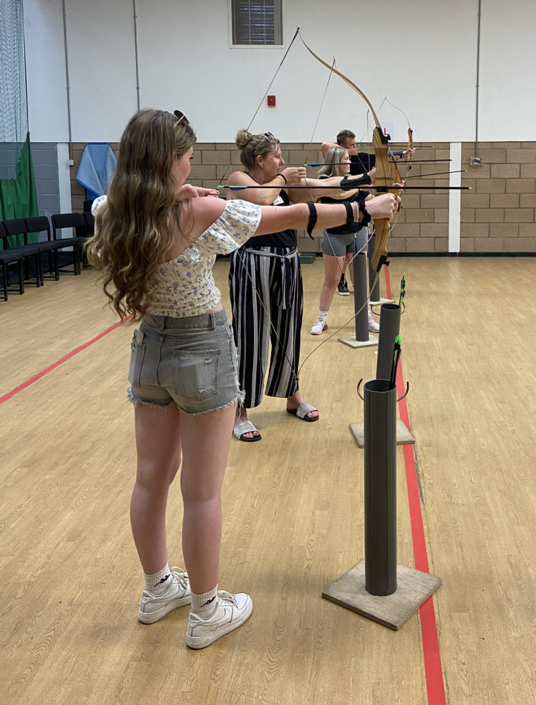 Four of the archers lined up in the indoor archery range, pulling their bows back ready to shoot their arrows at the targets.