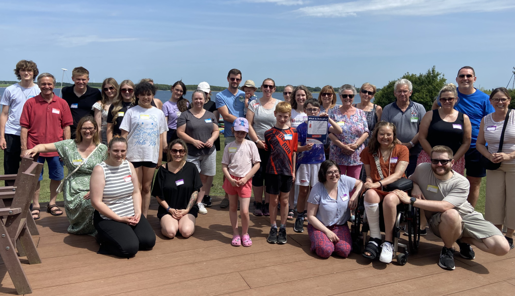 Group photo of the Behçet's friends and family day guests taken on a wooden deck with a blue sky and the tree-lined reservoir in the background.