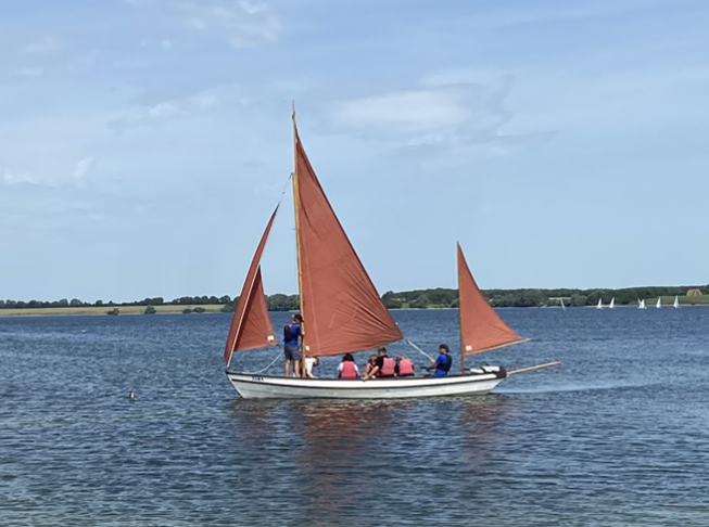 The white keel boat with three orange sails heading out to sail around the reservoir. The water is calm and the sky is blue with a few light clouds