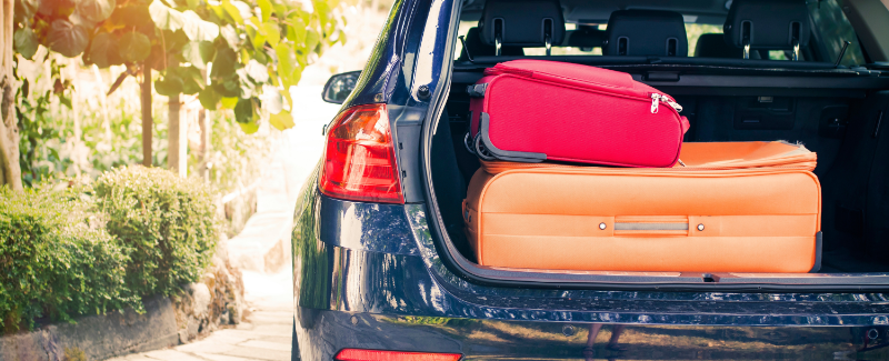An open car boot packed with a bright red suitcase stacked on top of a larger orange suitcase. The dark blue car is parked on a paved driveway next to lush green bushes and trees, with soft sunlight filtering through the leaves on the left.