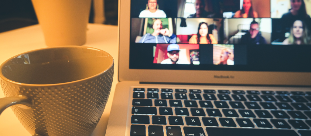 A close-up, slightly angled shot of a laptop screen displaying a video conference with multiple participants grid-style. A large, textured ceramic coffee mug sits in the sharp foreground on the left. The background is softly blurred, showing a table surface and a potted plant under warm, ambient lighting.
