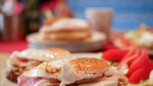 Close-up photo of a fried egg on top of a slice of bacon and some bread. Out of focus behind it are some tomatoes and another fried egg. There is another plate of food and a drink out of focus in the background.
