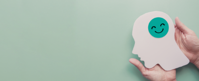 Hands gently holding a white paper silhouette of a human profile with a teal smiley face inside, set against a calm, solid light green background.